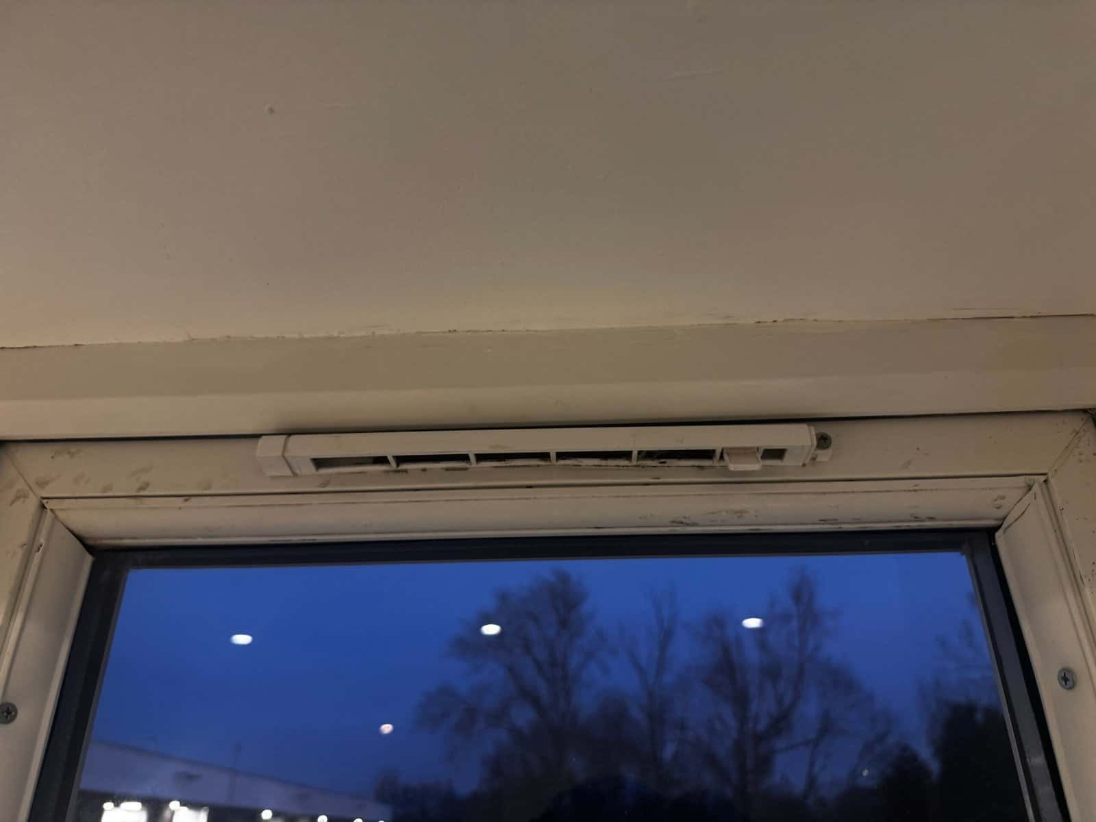 Top of a window in a Greenford home with a visible gap to the ceiling, a grimy trickle vent and failed sealant allowing cold air and damp in
