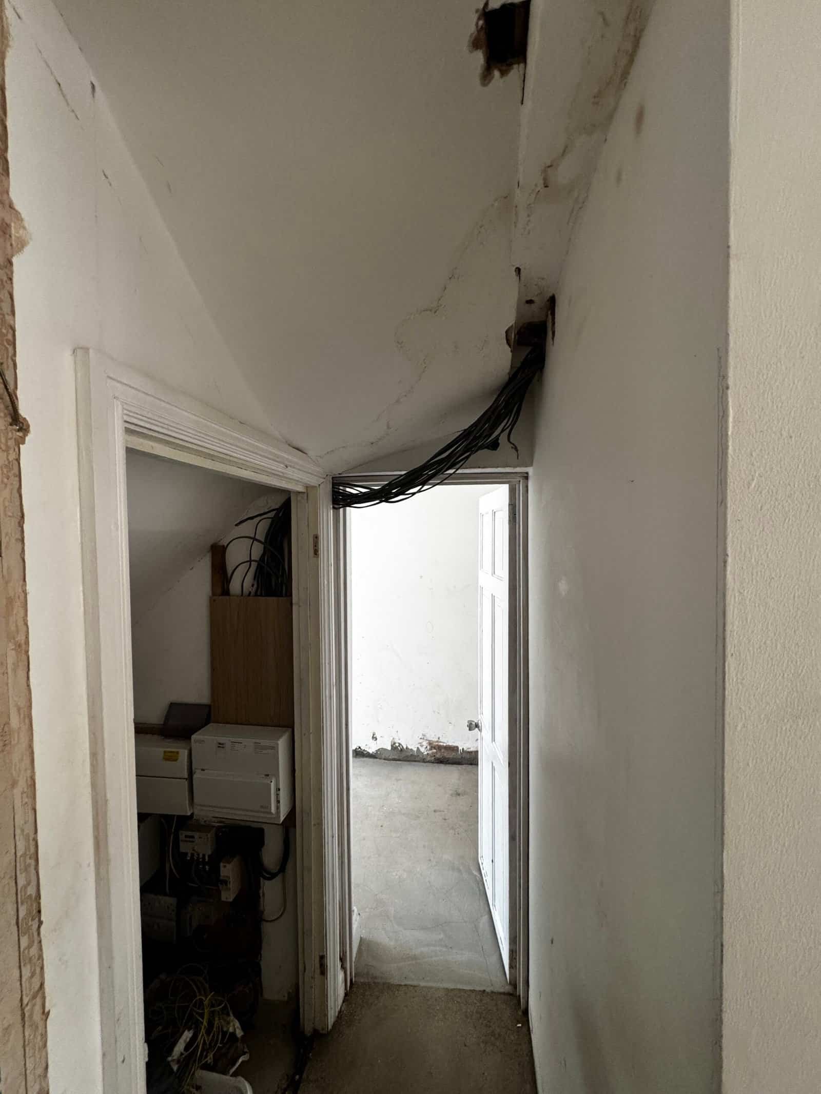 Hallway in a Greenford home with a hole knocked through the wall, loose cables draped across the ceiling and an exposed consumer unit and boiler cupboard
